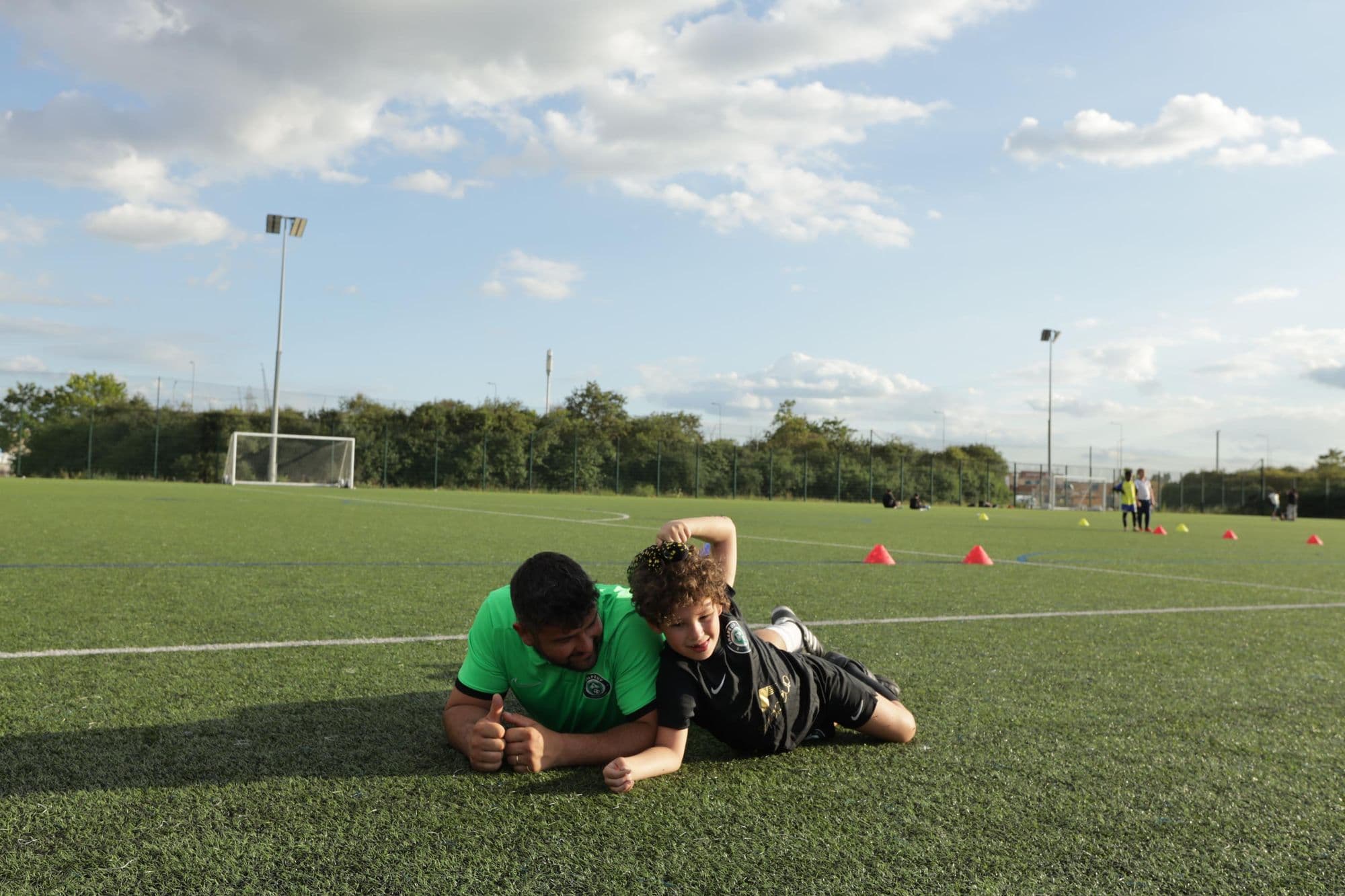 A coach and a young player lie on the pitch side by side, sharing a laugh