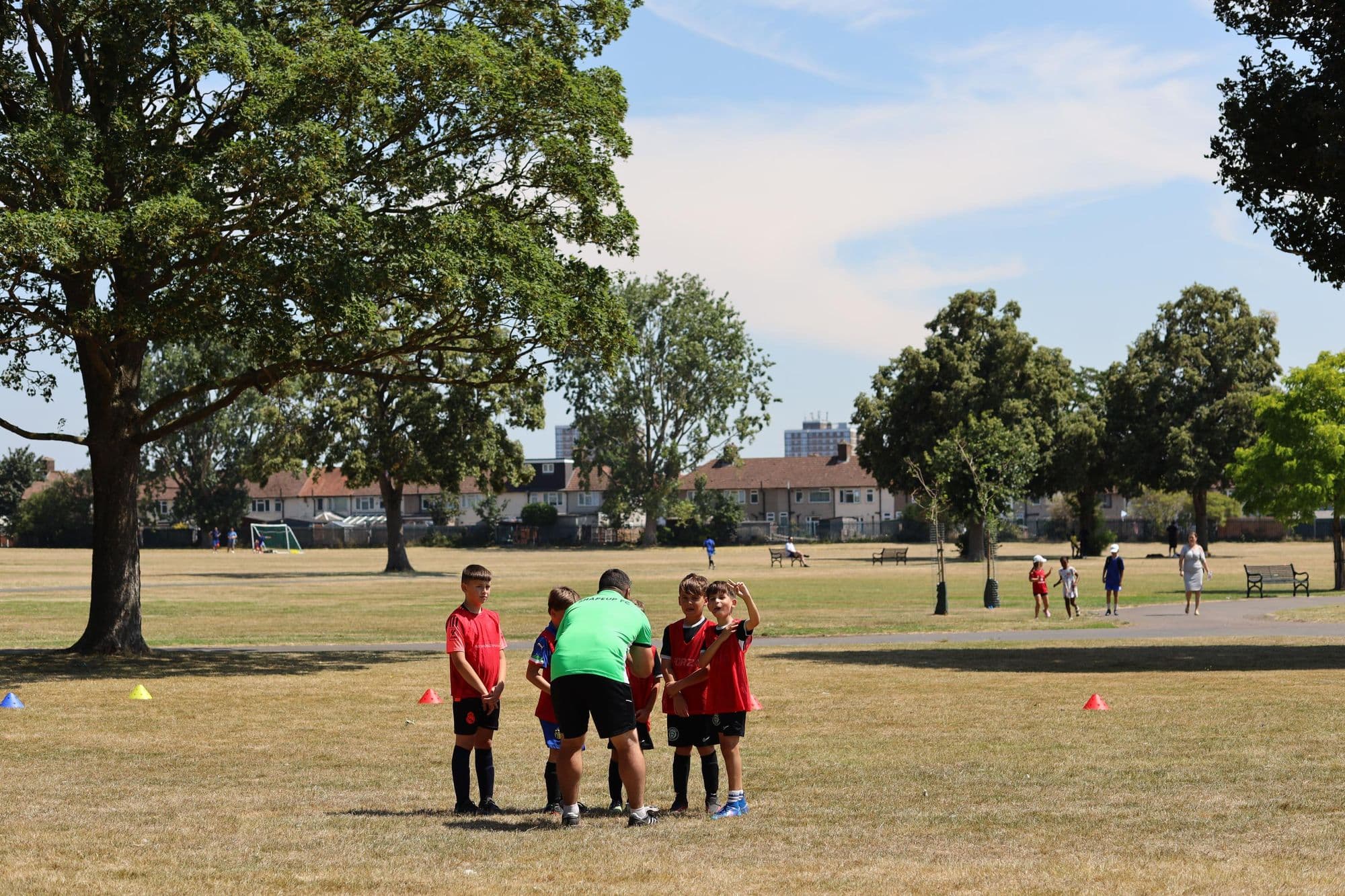 A coach crouches to talk with players in red bibs on a sunny day in the park